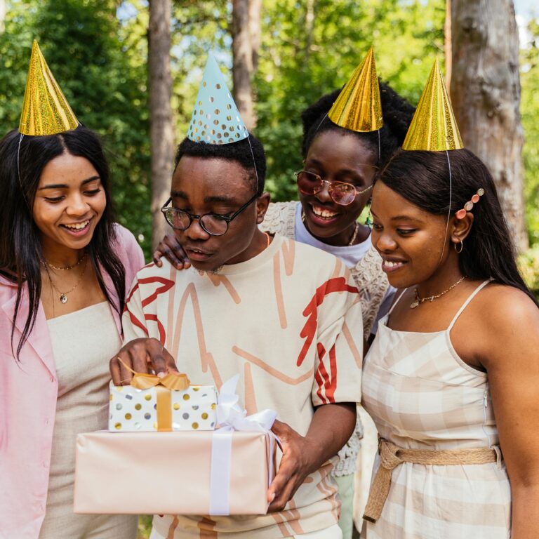 Group of friends celebrating a birthday outdoors with gifts and party hats, surrounded by nature.